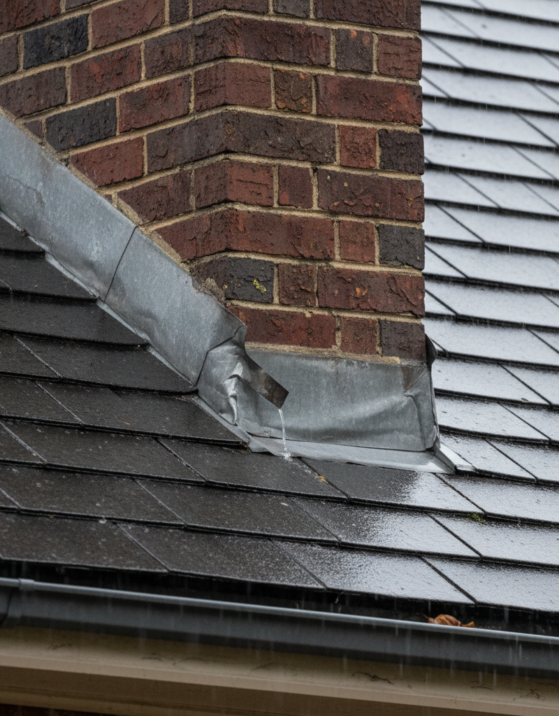 Leaking brick chimney where it meets a shingle roof, showing damaged flashing and a cracked crown during rain.