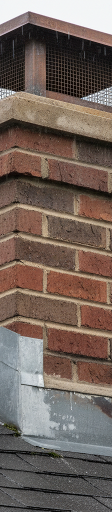 Close-up of a brick chimney meeting a shingled roof during rain, showing flashing