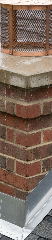 Close-up of a chimney on a residential roof during rain, showing water draining properly off the chimney cap and flashing, or pooling near a visible crack in the crown or lifted flashing.