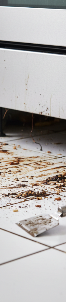 Kitchen floor with baked-on grease and food debris exposed under a pulled-out stove, with cleaning tools nearby.