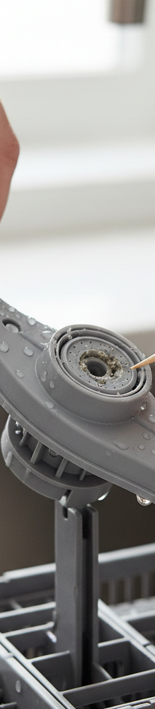 Hands cleaning a clogged dishwasher spray arm with a toothpick to remove debris.