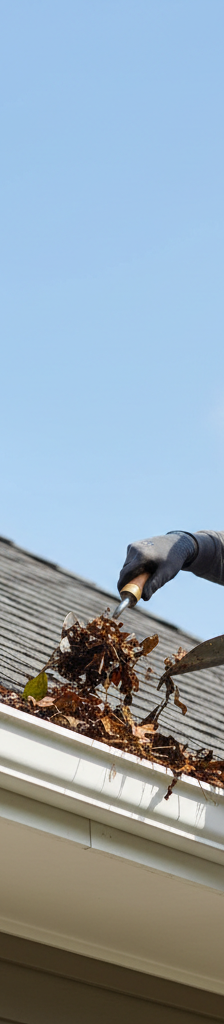 Homeowner on a ladder cleaning leaves and debris from a gutter to prevent roof damage
