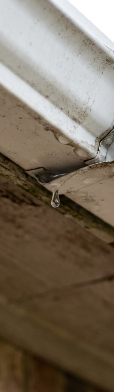 A close-up of a white gutter leaking at the seam, with a water droplet forming.