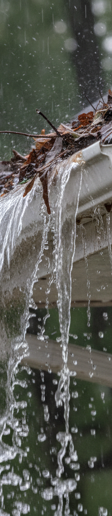 Gutter overflowing with water and leaves during a heavy rainstorm, indicating a clog or other issue.