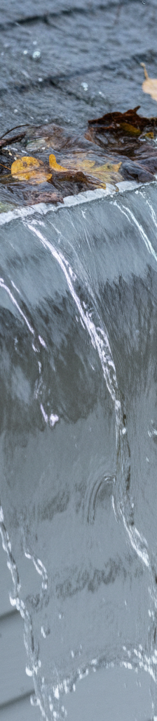 Gutter overflowing with water during a heavy rainstorm, with leaves and debris visible inside.