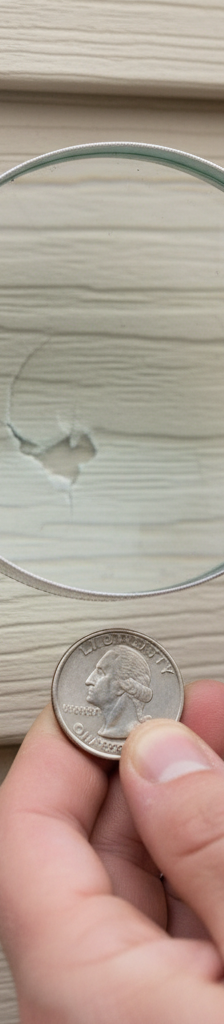 Close-up of vinyl siding with subtle crescent-shaped hail damage next to a magnifying glass.