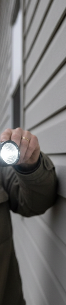 Homeowner inspecting vinyl siding for hail damage with a flashlight at an angle, looking for subtle dents and cracks.