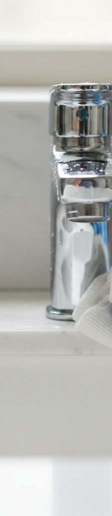 Close-up of a bathroom faucet half covered in hard water stains, half clean, being wiped by a gloved hand.