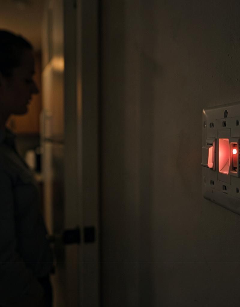 A homeowner in a dim hallway looking at an electrical panel with a tripped breaker glowing red