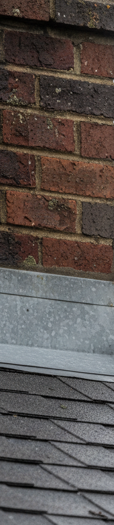 Close-up of storm-damaged metal flashing lifted from a brick chimney on a shingled roof, revealing a small gap.
