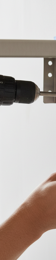 Close-up of a hand using a drill/driver to install a floating shelf bracket, with one screw driven into a wall stud and another hole ready for a toggle anchor in drywall.