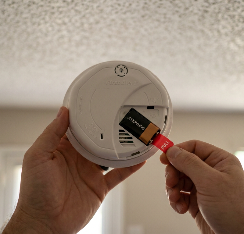 A person holding a new smoke detector with the battery compartment open, showing the unremoved plastic pull-tab.