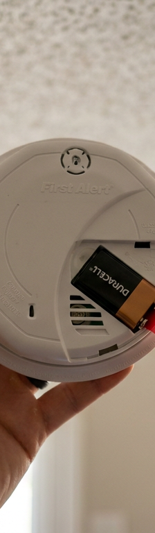 A person holding a new smoke detector with the battery compartment open, showing the unremoved plastic pull-tab.
