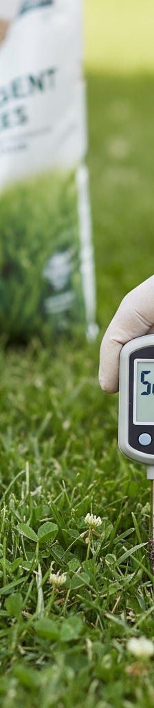 Hand holding a soil thermometer in a green lawn, showing 50-55°F reading near a bag of pre-emergent.