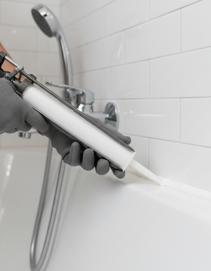 Person in gloves carefully applying a bead of fresh silicone caulk around a bathtub edge.