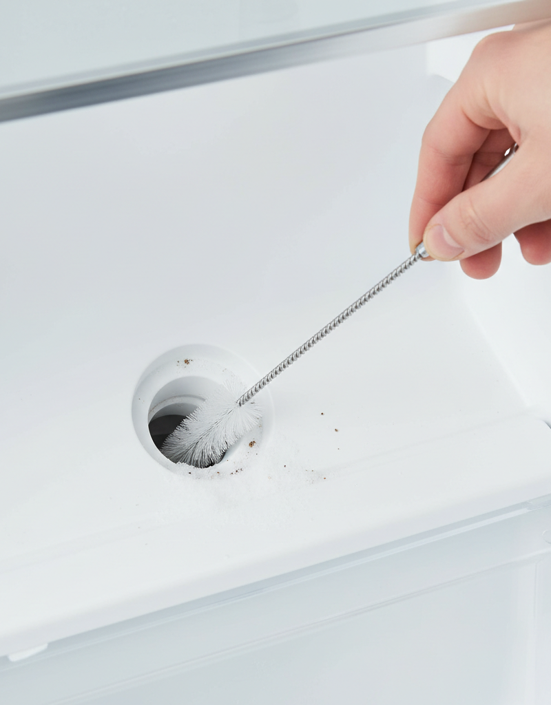 Hand cleaning a clogged refrigerator defrost drain tube with a flexible brush in a freezer compartment.