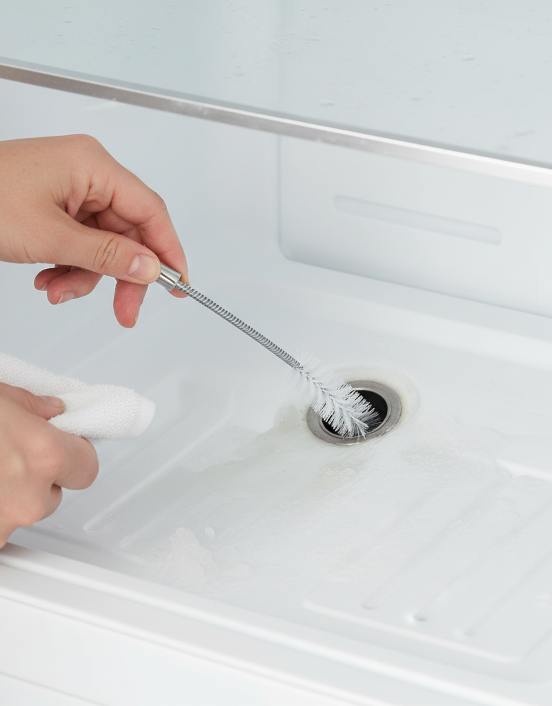 Homeowner using a thin brush to clear a clogged defrost drain in the freezer of a refrigerator, showing the interior of the freezer.