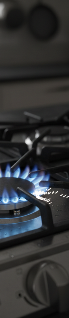 A close-up shot of a gas stove burner with the blue flame lit while the white ceramic igniter is visibly sparking next to it.