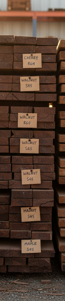 A stack of S2S walnut lumber with two smooth faces and rough edges is next to a stack of S4S poplar lumber, smooth on all four sides, illustrating the key difference for a woodworker.