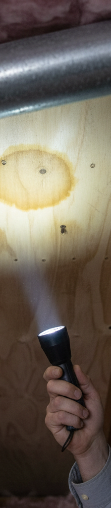 Homeowner inspecting an attic for early signs of a roof leak, shining a flashlight on a water stain.