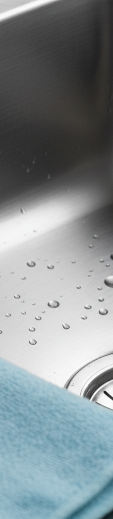 A perfectly polished, gleaming stainless steel kitchen sink with no haze or water spots, reflecting light, with water beads on the surface.