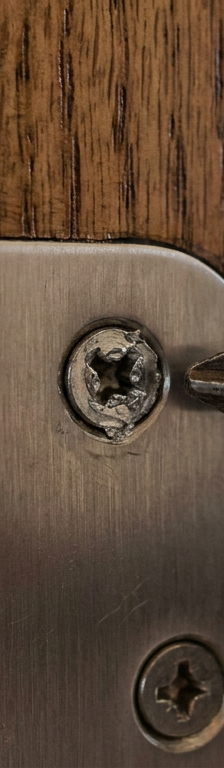 A close-up of a stripped screw in a door hinge, showing the damaged head that the drill can no longer grip.