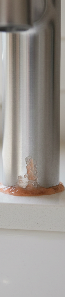 Close-up of pinkish-orange slime buildup around the base of a kitchen faucet on a light countertop.